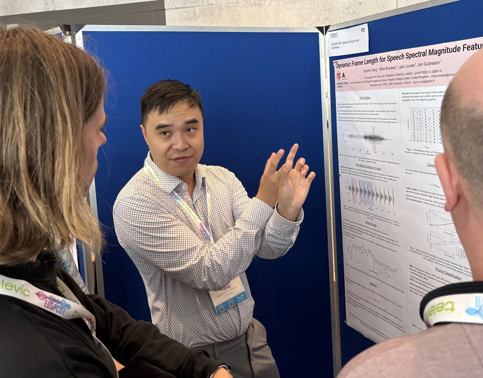 Guolin Fang mid-explanation, one hand raised, talking to two attendees at the EUSIPCO 2026 poster session; the attendees are seen from behind in the foreground. His poster, 'Dynamic Frame Length for Speech Spectral Magnitude Features', is on the display board to the right, showing the problem statement, waveform plots and time-frequency tiling diagrams.